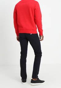 Felpa rossa con maniche lunghe, abbinata a pantaloni blu scuro e sneakers nere. La felpa ha orlo e polsini a costine.