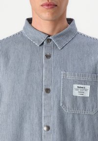 Camisa aos riscas em azul e branco, com colarinho, botões metálicos e um bolso com remendo que apresenta um rótulo. Textura suave, design clássico.