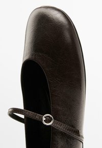 Brown leather Mary Jane shoe with rounded toe, featuring a thin strap and circular metal hardware, against a light background.
