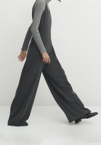 Combinaison grise à jambes larges portée sur un haut gris ajusté à manches longues. Tissu lisse et texturé ; lignes épurées ; design minimaliste ; chaussures noires.