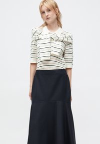 Young woman with blonde hair wearing a cream and black striped sweater tied over shoulders and a high-waisted black skirt.