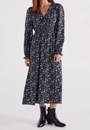 Femme portant une robe midi noire à manches longues avec un imprimé floral blanc et rose pâle, associée à des bottines noires.