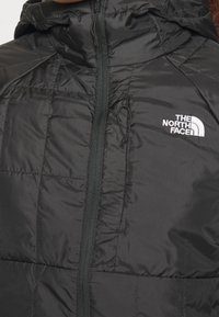 Svart isolerad jacka med dragkedja framtill och justerbar huva. Har kviltad textur och kontrasterande vit logotyp på bröstet.