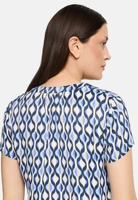 Blouse à manches courtes avec un motif de vagues répétées en bleu marine, bleu et beige. Tissu léger et fluide avec un col haut.