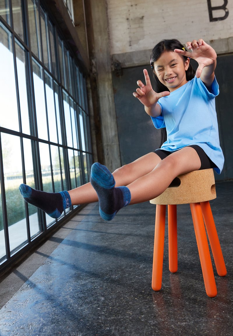 Korgistool oranžide jalgadega; laps sinises särgis ja tumedates lühikestes pükstes; sinine ja tume sokk; istub jalad rõõmsalt tõstetud.