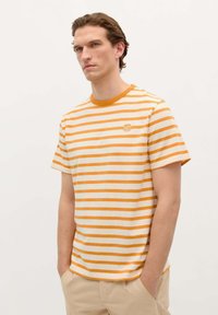 Camiseta a rayas en crema y naranja, con cuello redondo, mangas cortas y un pequeño logo en el lado izquierdo del pecho. Tejido de algodón suave.