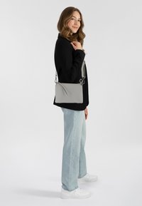 Donna con capelli mossi che indossa una giacca nera, jeans azzurri chiari, sneakers bianche e porta una borsa a tracolla grigia sulla spalla.