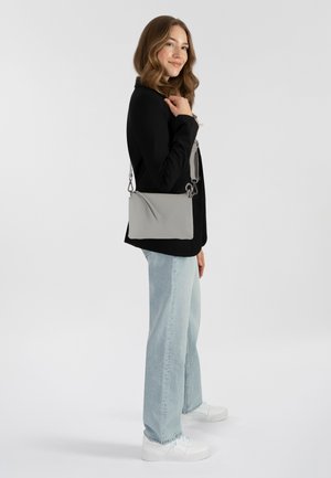 Femme aux cheveux ondulés portant une veste noire, un jean bleu clair, des baskets blanches et portant un sac en bandoulière gris sur son épaule.