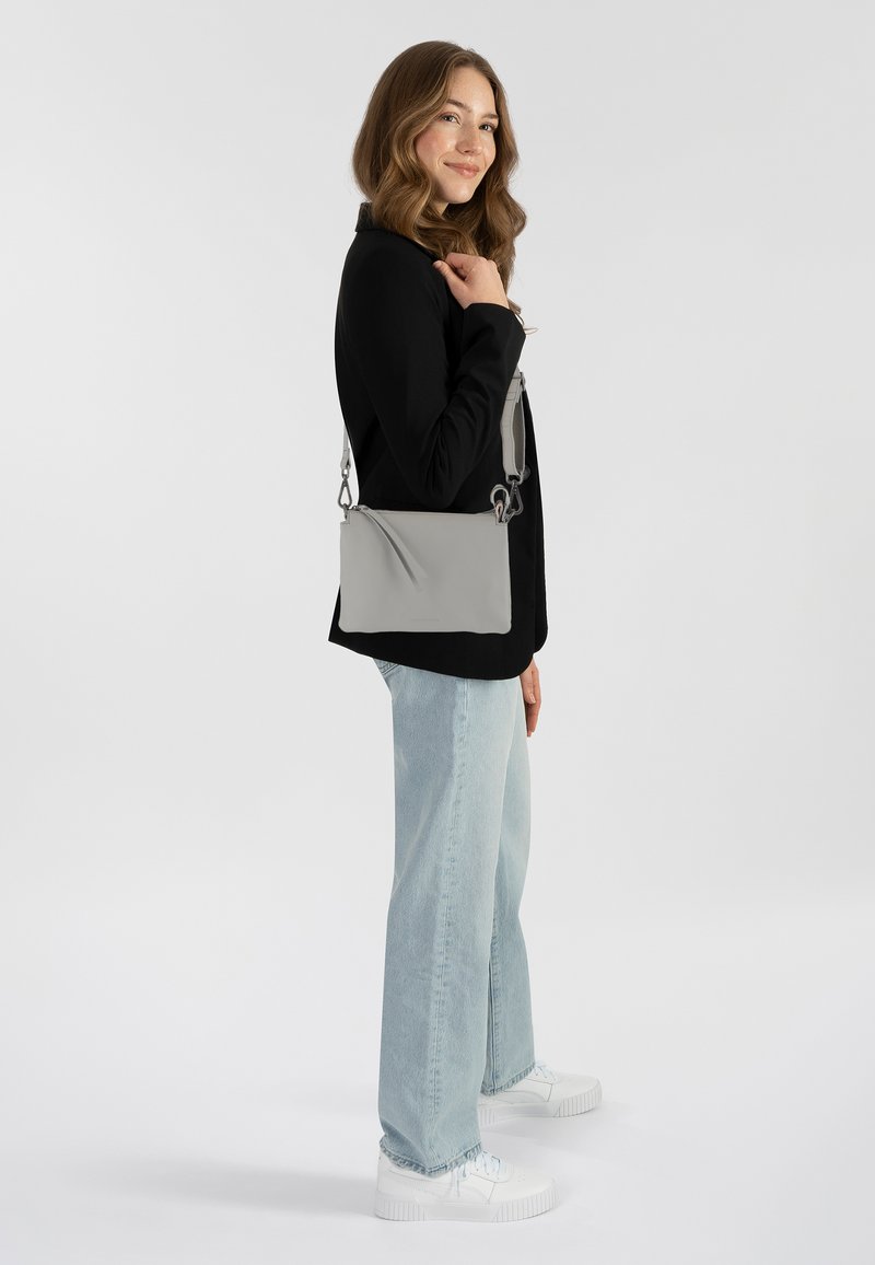 Donna con capelli mossi che indossa una giacca nera, jeans azzurri chiari, sneakers bianche e porta una borsa a tracolla grigia sulla spalla.