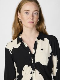 Žena s červenými vlasmi a svetlými pehami, oblečená v čiernej a béžovej košeli s kvetinovým vzorom a malými náušnicami, smeruje dopredu s neutrálnym výrazom.
