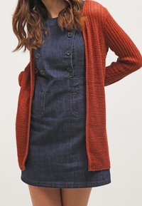 Cardigan lavorato a maglia di colore ruggine con apertura frontale e texture a coste, indossato sopra un vestito di jeans con bottoni e tasche applicate.