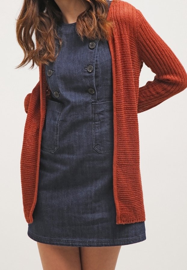 Cardigan lavorato a maglia di colore ruggine con apertura frontale e texture a coste, indossato sopra un vestito di jeans con bottoni e tasche applicate.