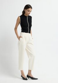 Zwarte mouwloze top met witte ritsdetails, gecombineerd met crèmekleurige geplooide high-waist broeken en zwarte puntige hakken. Glad textuur van de stof.