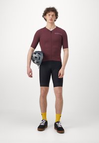 Burgunderfarbener Radtrikot mit kurzen Ärmeln und Reißverschluss vorne, kombiniert mit schwarzen Shorts. Legere schwarze Schuhe und hellgelbe Socken.