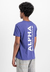 Lila pamut póló, a hátsó részén kiemelkedő fehér "ALPHA INDUSTRIES" grafikával. Rövid ujjú és kerek nyakú.
