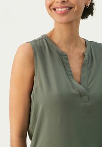 Top fără mâneci de culoare verde măslin, cu decolteu în V și detalii fine de cusătură. Material neted, croială lejeră, potrivit pentru ținute casual.