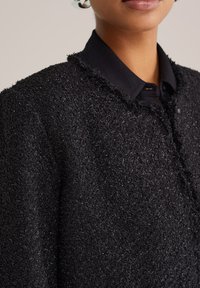 Suurel plaanil on inimene, kes kannab tekstuurset musta jakki musta kraeülikonna kohal, rõhutades kangaste üksikasju ja krae kuju.