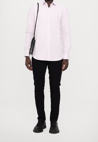 Chemise à rayures roses claires à manches longues, pantalon slim noir, chaussures noires et petit sac à bandoulière noir. La chemise a un col classique.