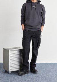 Grå huvtröja med framficka; svarta cargobyxor med flera fickor; svarta kängor; silverfärgad resväska på hjul bredvid modellen; neutral bakgrund.