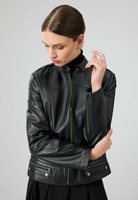 Zwarte leren jas met een hoge kraag, met rits afgesloten zakken, aansluitende snit en een gladde glans. Bevat metalen hardware accenten.