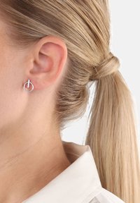 Orecchino a cerchio argentato con dettaglio a barra verticale indossato sull'orecchio sinistro, capelli biondi legati in una coda di cavallo, camicia bianca con colletto visibile.