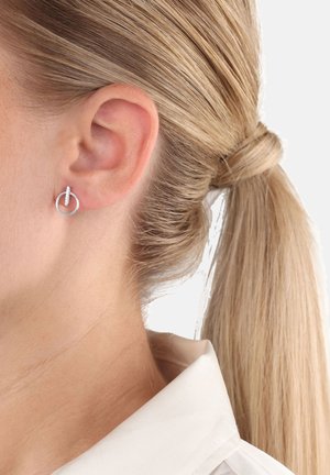 Orecchino a cerchio argentato con dettaglio a barra verticale indossato sull'orecchio sinistro, capelli biondi legati in una coda di cavallo, camicia bianca con colletto visibile.