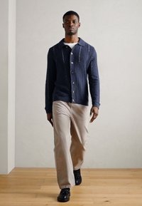 Sötétkék kötött kardigán bonyolult varrásmintákkal, gombos elülső résszel és széles gallérral; világos bézs nadrággal és fekete cipővel párosítva.