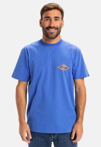 Blaues Baumwoll-T-Shirt mit rundem Halsausschnitt und kurzen Ärmeln. Es hat ein buntes, diamantförmiges Grafikdesign auf der linken Brustseite.