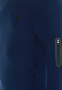 Tummansininen pitkähihainen paita, jossa on tekstuuri pinta, etuosassa puolivetoketju ja varmasti suljettava vetoketjullinen tasku vasemmassa hihassa. Hämähäkkilogo rinnassa.