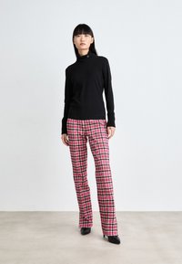 Schwarzer langärmeliger Rollkragenpullover mit einem dekorativen Knopf, kombiniert mit rosa und schwarzen Hahnentritt-Hosen und schwarzen High Heels.