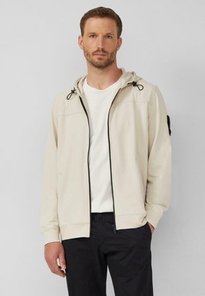 Lichtbeige zip-up hoodie met een trekkoordcapuchon, zijzakken en een contrasterende zwarte rits. Draag het over een wit shirt, gecombineerd met donkere broeken.
