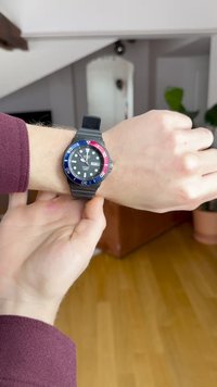 Črna gumijasta ura na zapestju z modrim in rdečim obročem, črn obraz s prikazom datuma in dneva. Kazalci in številčnice so bele. Klasičen dizajn.