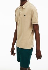 Polo beige hecho de tejido texturizado, con un logo bordado en verde, combinado con pantalones cortos verde oscuro, que muestran un ajuste relajado y aberturas laterales.
