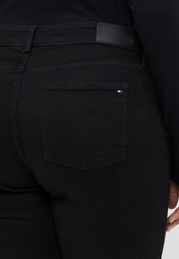 Black denim jeans with a smooth texture, featuring a back pocket and a leather label embossed with "TOMMY HILFIGER" at the waistband.