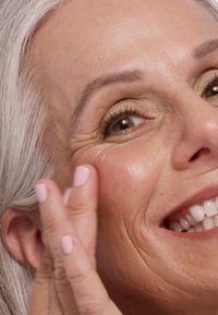 Primo piano di una persona che applica crema sulla guancia. Presenta pelle chiara, linee sottili, capelli grigi e smalto per unghie rosa chiaro.