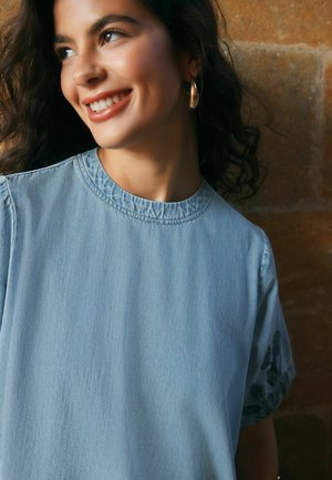 Junge Frau mit lockigem dunklem Haar lächelt, trägt goldene Creolen und ein hellblaues Jeansoberteil mit Rundhalsausschnitt vor einer Ziegelsteinwand.