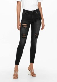 Jeans negros ajustados con parches desgastados. Presentan una cintura alta y dobladillos desflecados, combinados con sandalias de tacón transparente.