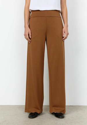 Vrouw draagt hoge taille, wijde bruine broek, wit topje en zwarte ballerina's, staand op een lichtgrijze vloer tegen een witte muur.