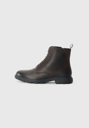 Botas de tobillo de cuero marrón con punta redondeada, aseguradas con seis ojales y una suela de goma negra. Con un tirador en la parte posterior.