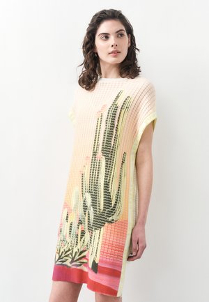 Frau trägt ein kurzärmliges, gestricktes Kleid mit Muster aus hohen Kakteen und Wüstenpflanzen in Grün-, Pink- und Rottönen auf hellem Hintergrund.