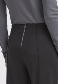 Zbliżenie na osobę noszącą czarne spodnie z tylnym zamkiem błyskawicznym oraz szary top z długim rękawem, pokazujące tors i górną część nóg.