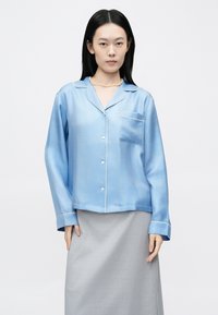 Világoskék szatén pizsama felső bemetszett gallérral, elülső zsebbel és fehér szegéllyel. Hosszú ujjú, laza fazon, szürke szoknyával párosítva.