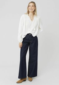 Blouse blanche à manches longues avec un tissu texturé et des détails à franges, associée à un jean embelli large en denim sombre à taille haute et des bottines châtaigne.