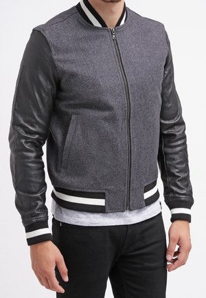 Mann trägt eine grau-schwarze Collegejacke mit Lederärmeln, gestreiften Bündchen und Bund über einem hellgrauen Hemd und schwarzen Hosen.