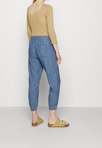 Vrouw in een tan lange mouw top, blauwe elastische taillebroek met achterzakken, en tan gespsandalen, staand tegen een effen achtergrond.