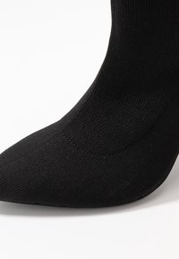 Černá ponožka s ribbed designem a špičatou špičkou. Látka vypadá pružně a texturovaně, pokrývá dolní část nohy. Bez viditelných vzorů nebo akcentů.