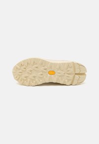Beige tursko med såle med en robust mønster og et gult Vibram-logo; laget av gummi for bedre grep og holdbarhet.