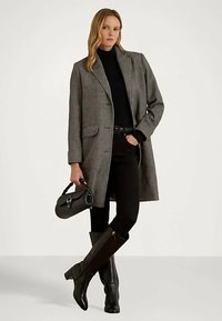 Sieviete, tērpusies pelēkā garā mētelī, melnā turtleneck, melnās biksēs un melnās augstpapēžu zābakos līdz ceļgaliem, tur mazu melnu rokassomu.