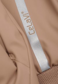 Beige vattentätt tyg med en glänsande silveraccentetikett. Tyget är slätt, med en strukturerad stickad mudd i botten.