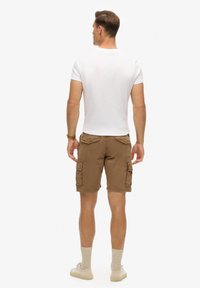 Brune cargo shorts med klaplommer, parret med en hvid T-shirt med rund hals. Modellen har beige sokker og lyse sko på.